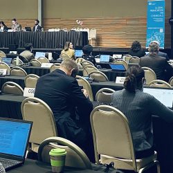 People sit at rows of tables with laptops and documents during a conference or meeting. A panel of speakers is seated at a long table at the front of the room. A large screen displays text and graphics.