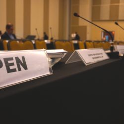 A row of tables with nameplates and microphones in a conference room. The nameplate in the foreground reads IPEN. Several empty chairs and a few people sitting in the background are visible.