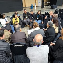A group of people sit in a circle having a discussion in a large indoor hall. Rows of empty black chairs surround them, and some tables and equipment are visible in the background.