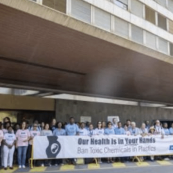 A group of people stand in front of a large building holding a long banner that reads, Our Health is in Your Hands. Ban Toxic Chemicals in Plastics. The group appears to be participating in a demonstration.