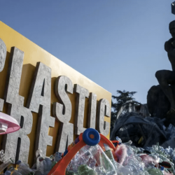 一个大大的黄色牌子，上面写着“PLASTIC”，周围堆满了塑料垃圾。背景中，一个酷似罗丹《思想者》的雕像坐落在更多的塑料上，其后是一个DNA双螺旋结构。.