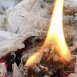 A close-up view of a small flame burning on a pile of crumpled paper and debris. The background appears out of focus.