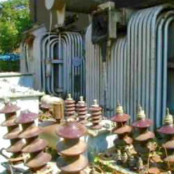 An outdoor electrical substation with several large ceramic insulators, metal structures, and coiled pipes mounted on a building wall. The area is exposed to sunlight and partially shaded.