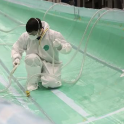 A person wearing a white protective suit and face mask is kneeling inside a large, empty, turquoise industrial tank while handling hoses.
