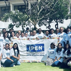 A group of people in matching IPEN t-shirts pose outdoors, holding a large banner that reads “IPEN for a toxics-free future IPEN South Asia,” with a building and trees in the background.