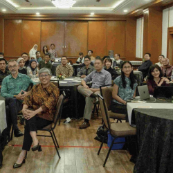 Një grup njerëzish ulen rreth tavolinave në një dhomë konferencash, duke u përballur me kamerën. Disa buzëqeshin, ndërsa të tjerë duken neutralë. Dhoma ka mure me panele druri dhe një dritë të madhe në tavan. Laptopët dhe letrat janë mbi tavolina.