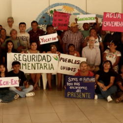 A large group of people stand indoors holding colorful signs with various messages, including “Seguridad Alimentaria,” “Pueblos Unidos,” and “Quintana Roo,” with a mural in the background.