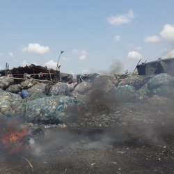 Large piles of plastic waste are stacked outdoors near makeshift structures. Smoke and flames rise from burning debris in the foreground, under a blue sky with scattered clouds.