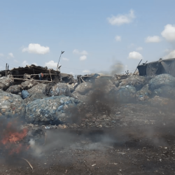 Large piles of plastic waste are stacked outdoors near makeshift structures. Smoke and flames rise from burning debris in the foreground, under a blue sky with scattered clouds.