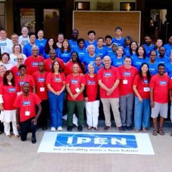 A large group of people pose for a photo outside a building, wearing red, blue, yellow, or white IPEN T-shirts. A banner on the ground reads IPEN for a healthy, toxics-free future.
