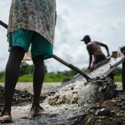 Two people work outdoors using tools to sift and wash gravel in shallow water. One person stands close, seen from behind holding a shovel, while the other is further away operating a sluice. The sky is cloudy.
