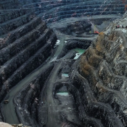 A large open-pit mine with stepped, terraced sides, winding roads, and several pools of water at the bottom. Machinery and vehicles are visible on different levels of the excavation.