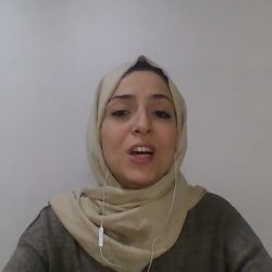A woman wearing a beige hijab and a dark top is speaking. She has a wired earbud in her left ear and is seated in front of a plain white wall.